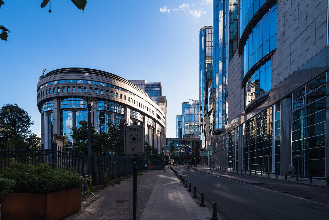 The curved glass Paul-Henri Spaak building of the European Parliament in Brussels, next to modern office towers under a blue sky.